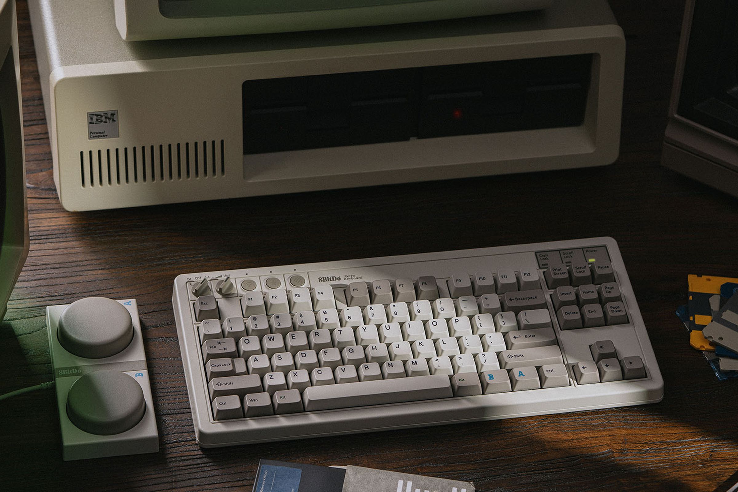 Keyboard mekanis 8BitDo Edisi M di atas meja di samping tombol makronya, di depan komputer IBM vintage.
